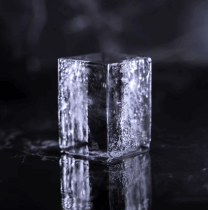 Un glaçon transparent et rectangulaire se tient debout sur une surface sombre et réfléchissante, où l'on distingue une légère condensation et une réfraction de la lumière.