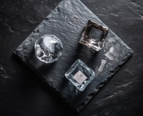 Trois blocs de glace, un sphérique et deux cubiques, posés sur une plaque de pierre noire avec des étiquettes distinctes, créant une composition minimaliste et élégante.
